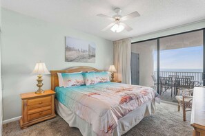 A bedroom with a bed, two bedside tables, a ceiling fan, a painting, and a view of the sea through a balcony door. - Pelican Pointe1204: 3BR/ 2BA, gulf-front. (Orange Beach)