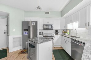 A modern kitchen with white cabinets, stainless steel appliances, a granite countertop, and a green rug. - Pelican Pointe1204: 3BR/ 2BA, gulf-front. (Orange Beach)