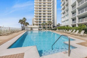A swimming pool with a clear blue water, surrounded by lounge chairs and palm trees, with a tall building in the background. - Pelican Pointe1204: 3BR/ 2BA, gulf-front. (Orange Beach)