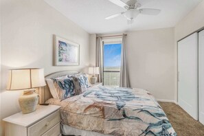 A bedroom with a bed, a nightstand, a lamp, a ceiling fan, a window with blinds, and a door. - Pelican Pointe1204: 3BR/ 2BA, gulf-front. (Orange Beach)