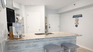 A kitchen with a granite countertop, a water dispenser, a sink, and two bar stools. - Harbour Place 102-  gulf-view, out & indoor pool, hot tub (Orange Beach)