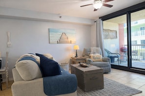 A living room with a sofa, armchair, and a small table with a lamp. There is a painting on the wall and a view of the outside balcony.