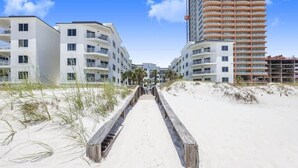 A beachfront walkway leading to a modern hotel with multiple balconies and a beachside view. - Palm Beach 22-D, beachfront, beach-view, 2nd floor (Orange Beach)