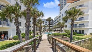 A wooden boardwalk leading to a pool area surrounded by palm trees and modern buildings. - Palm Beach 22-D, beachfront, beach-view, 2nd floor (Orange Beach)