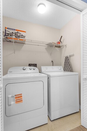 A laundry room with two white Whirlpool washers and dryers, a shelf with a TRAMONTINA appliance box, and a towel rack with a patterned towel. - Sea Oats 102C- Beach view, Pool, 25 steps to beach (Gulf Shores)