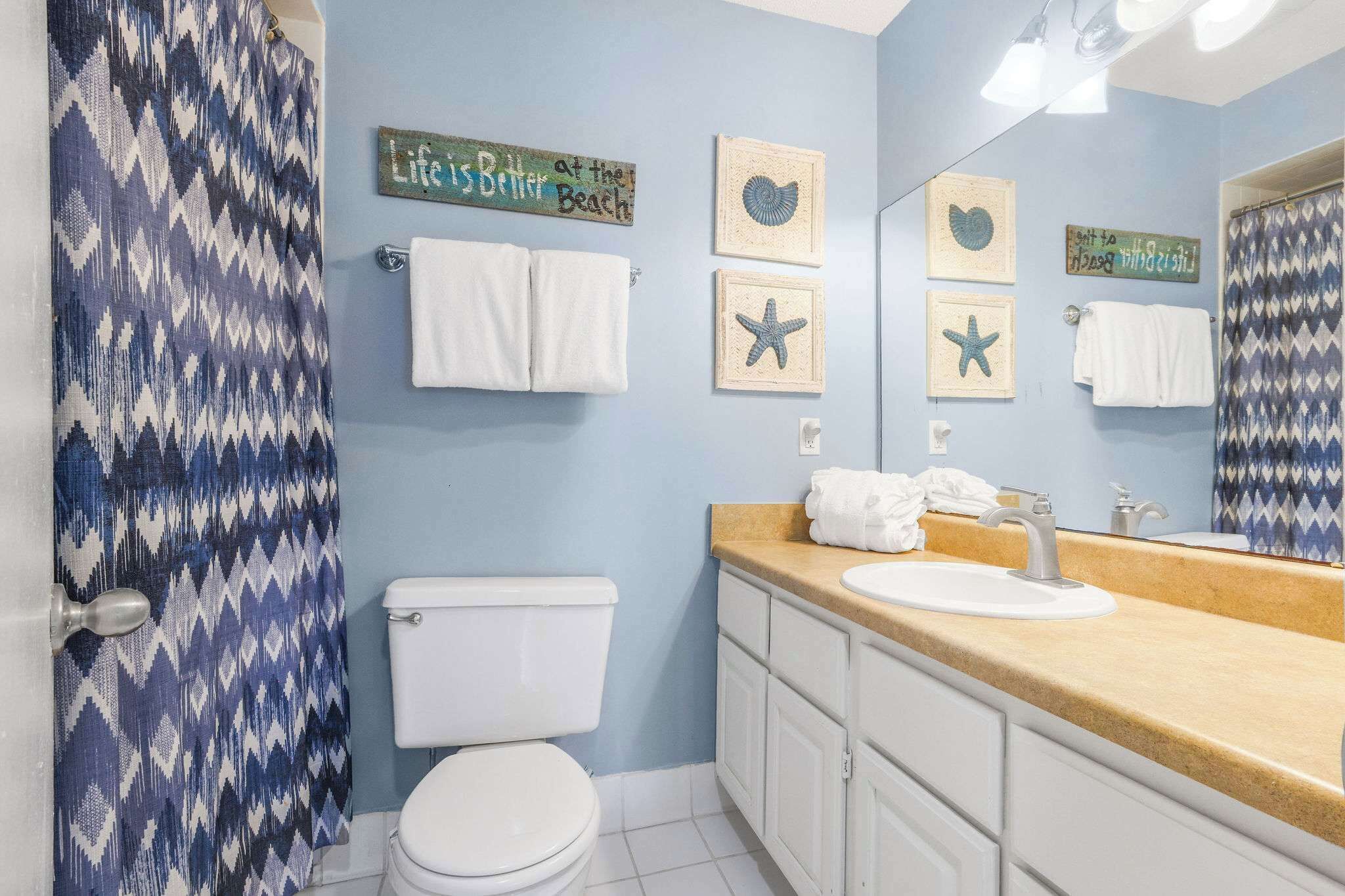 Un baño con cortina de ducha azul y blanca estampada, un inodoro blanco, un lavamanos con encimera color beige y adornos de estrellas de mar colgados en la pared.