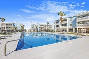A swimming pool surrounded by multi-story buildings with balconies, palm trees, and a clear sky.