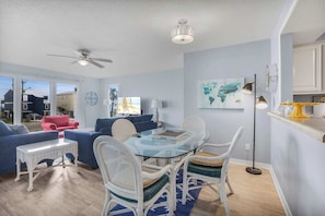 A modern living room with a glass-top dining table, white wicker chairs, a red armchair, and a ceiling fan.