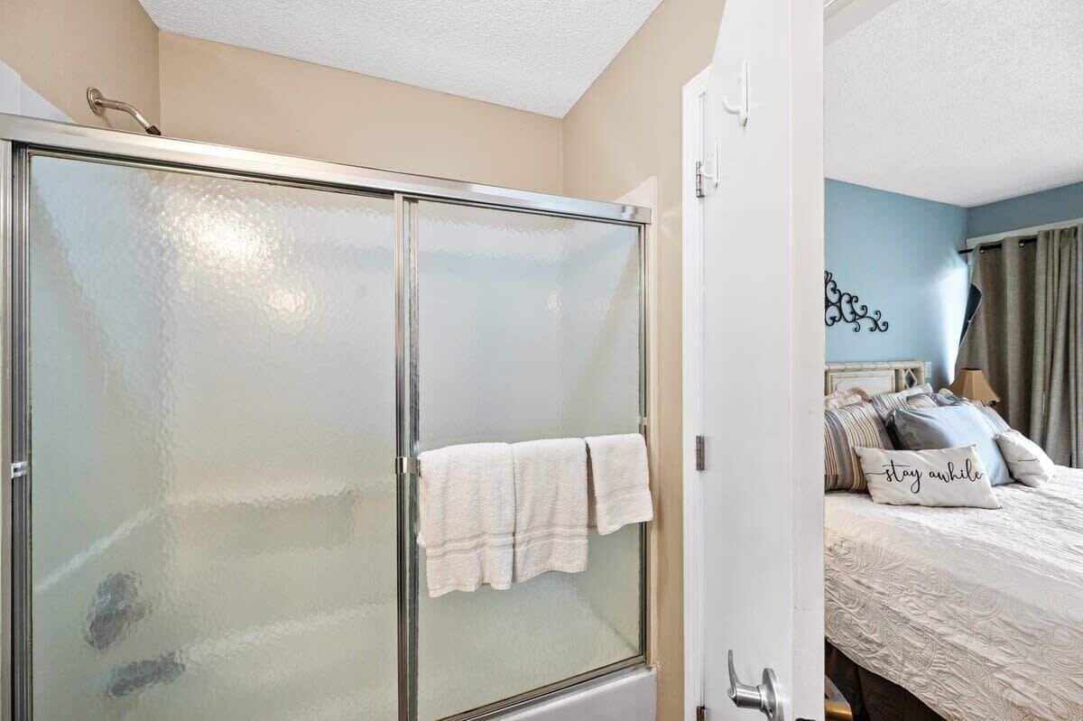 A bathroom with a glass shower enclosure and a bed with a 'stay awhile' pillow.