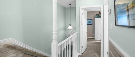 A hallway with a white railing, a hanging light fixture, and a door leading to another room.