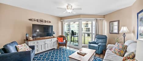 A living room with a flat-screen TV, a ceiling fan, a large window, and a blue patterned rug.