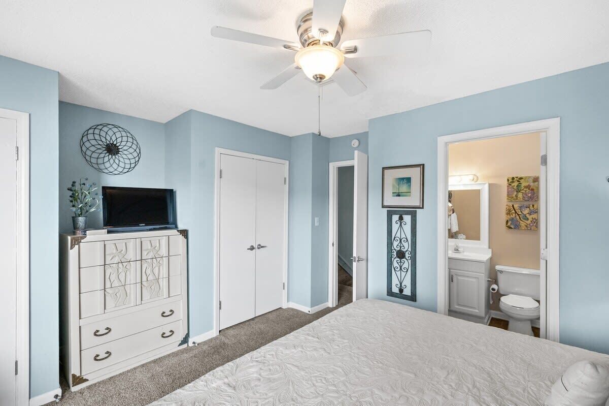 A bedroom with a bed, a dresser, a ceiling fan, and a door leading to a bathroom.