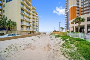 A beachfront hotel with multiple balconies, a sandy path leading to the beach, and palm trees.