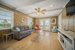 A room with a wooden ceiling, a ceiling fan, a gray sofa, a small table, a colorful ottoman, and a TV on a cabinet. - Don't Worry, Beach Happy; 6 BR, 4 BA, sleeps 20. (Gulf Shores)