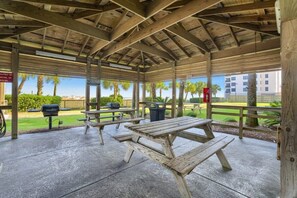 A covered picnic area with wooden benches and picnic tables, a barbecue grill, and trash cans.