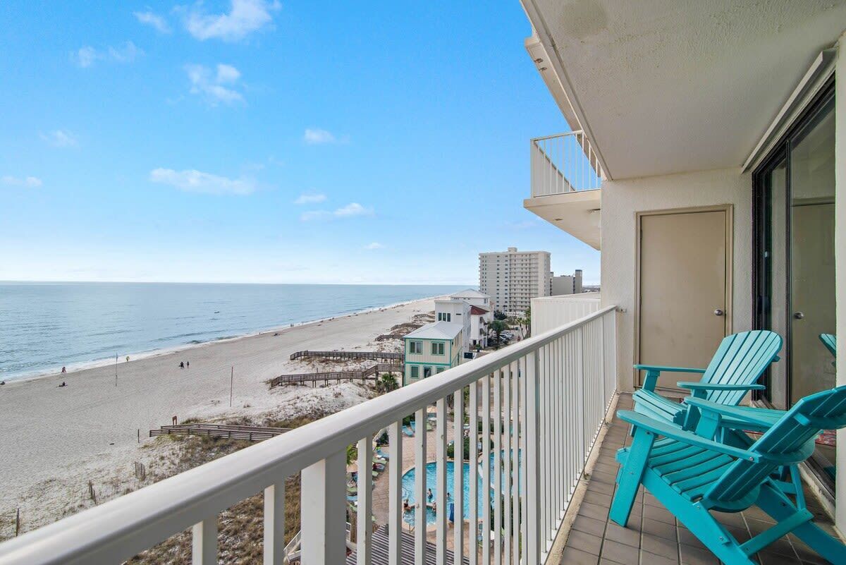 A balcony with a view of the beach, a pool, and a building in the background.