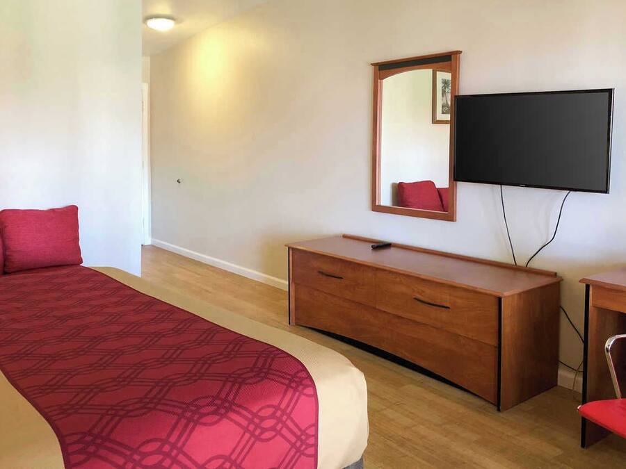 A hotel room with a bed, a wooden dresser, a desk with a chair, and a flat-screen TV mounted on the wall.