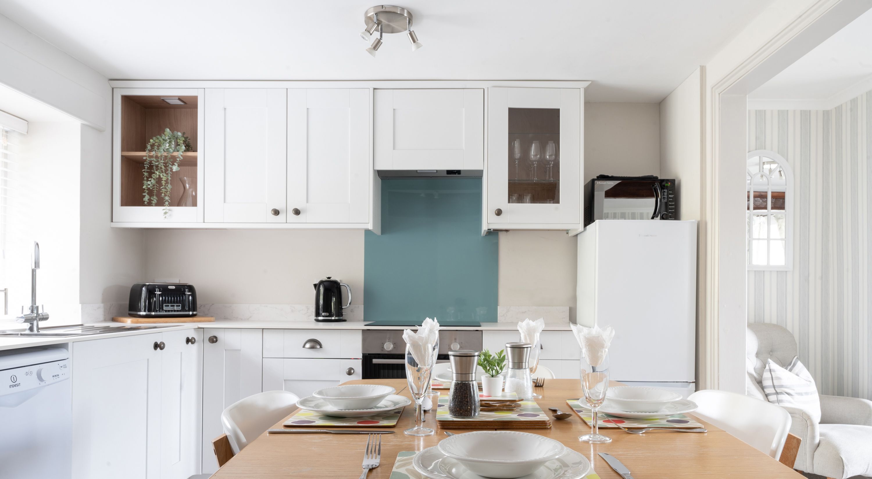 Une cuisine moderne avec des meubles blancs, un pare-feu turquoise et un coin repas aménagé pour quatre personnes, avec de l’assiette et des couverts blancs.