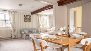 Een eetgedeelte met een houten tafel voor vier personen, witte stoelen en een gezellige zithoek met een bank en een raam met gordijnen.
