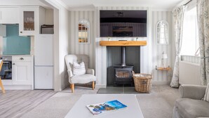 Een gezellige woonkamer met een open haard, een comfortabele fauteuil, een salontafel met een tijdschrift, en een keukenhoek met een turquoise apparaat.