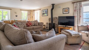 A cozy living room with a sofa, ottoman, and a television. There's a fireplace and a stone wall. - Stunning Lake fronted Property with Hot Tub - Hammerhole (Cunsey, Graythwaite)
