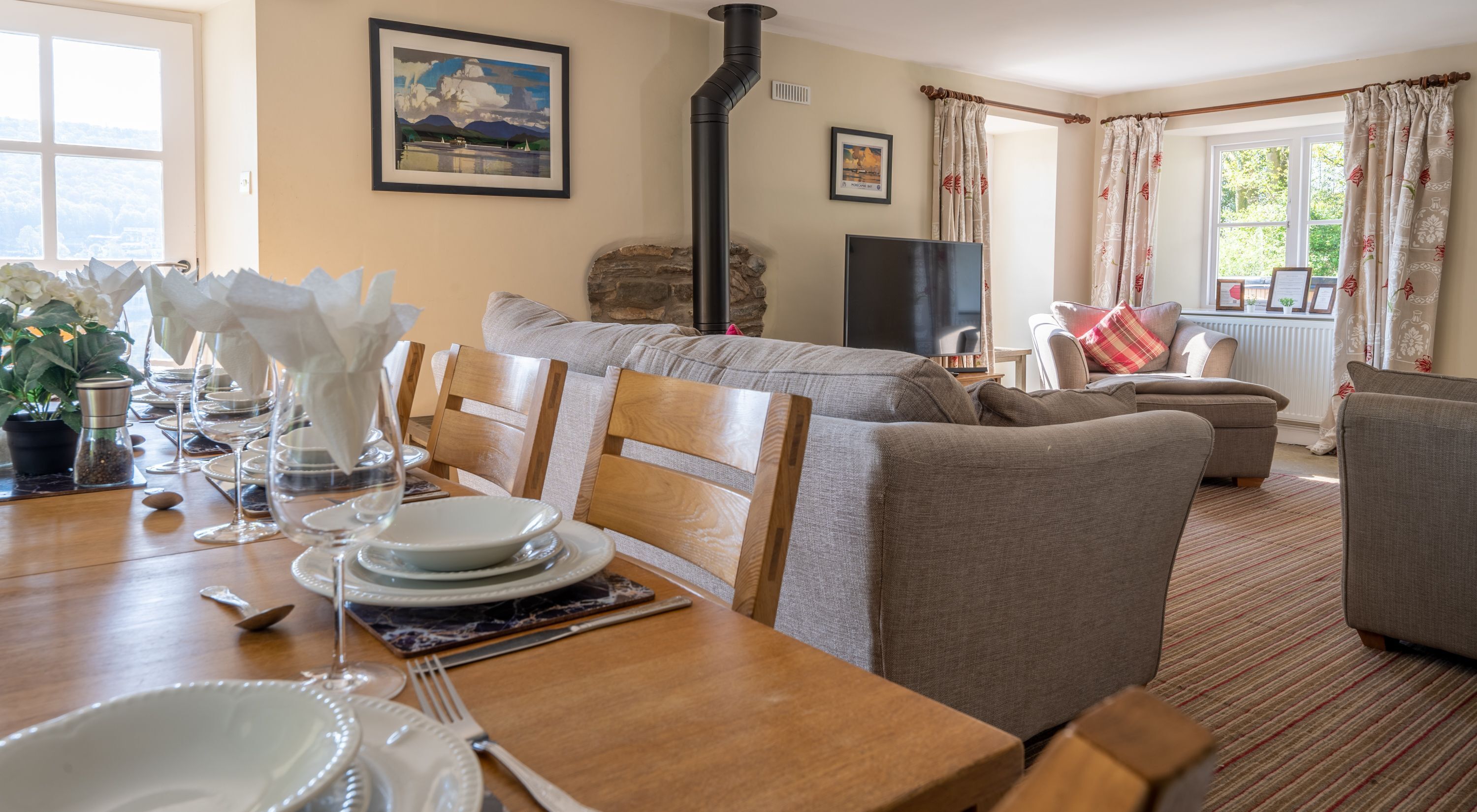A dining area with a wooden table set for a meal, a stone fireplace, and a living room with sofas and a television.