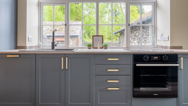A modern kitchen with grey cabinets, a built-in oven, and a window overlooking a garden.