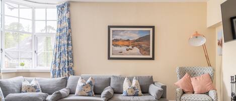 A living room with a grey sofa, patterned cushions, a wooden coffee table, a framed landscape painting, a patterned armchair, and a floor lamp.