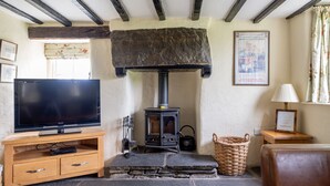 A cozy living room with a stone fireplace, a television on a wooden stand, and a wicker basket. - Lakeshore cottage with Hot Tub - Hullet Hall (Graythwaite)