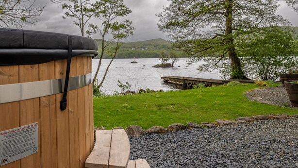 A wooden hot tub with a metal frame and a warning label, situated by a lake with a dock and trees in the background. - Lakeshore cottage with Hot Tub - Hullet Hall (Graythwaite)