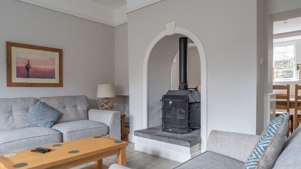 A living room with a grey sofa, a wooden coffee table, and a black wood-burning stove. - Fishery Views - Keepers Cottage (Graythwaite)