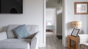 A living room with a grey sofa, a patterned pillow, a framed picture on the wall, and a small table with a lamp. - Fishery Views - Keepers Cottage (Graythwaite)