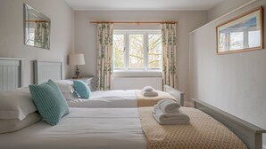 A hotel room with two beds, a bedside table with a lamp, a window with curtains, and a painting on the wall. - Fishery Views - Keepers Cottage (Graythwaite)