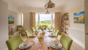 A dining room with a long table set for a meal, green chairs, and a view of the countryside through large windows.