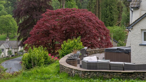 A stone wall with outdoor seating, a hot tub, and a large tree with red leaves.
