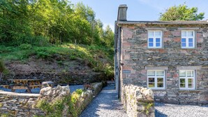 Ein Steinhhaus mit einem Kiesweg, der zum Eingang führt, umgeben von Grünflächen und einer Steinmauer.