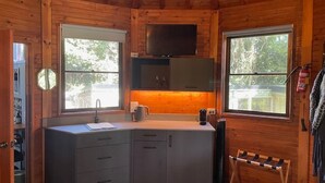 Cocina pequeña y funcional, con lavamanos, encimera y alacenas. Dos ventanas ofrecen luz natural y vistas al exterior.
