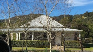 A white building with a peaked roof, surrounded by trees and a well-maintained hedge. - Lastover Cottage - Kangaroo Valley (Wattamolla)