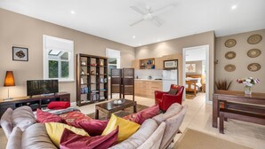 A modern living room with a sofa, TV, bookshelf, and a kitchen in the background. - Lastover Cottage - Kangaroo Valley (Wattamolla)