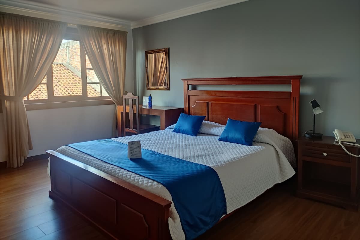 A neatly arranged hotel room with a large bed, a wooden nightstand, a desk, and a window with curtains.