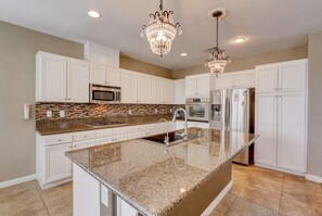 Uma cozinha moderna com armários brancos, bancadas de granito, uma ilha central, eletrodomésticos de aço inoxidável e duas luminárias de cristal.