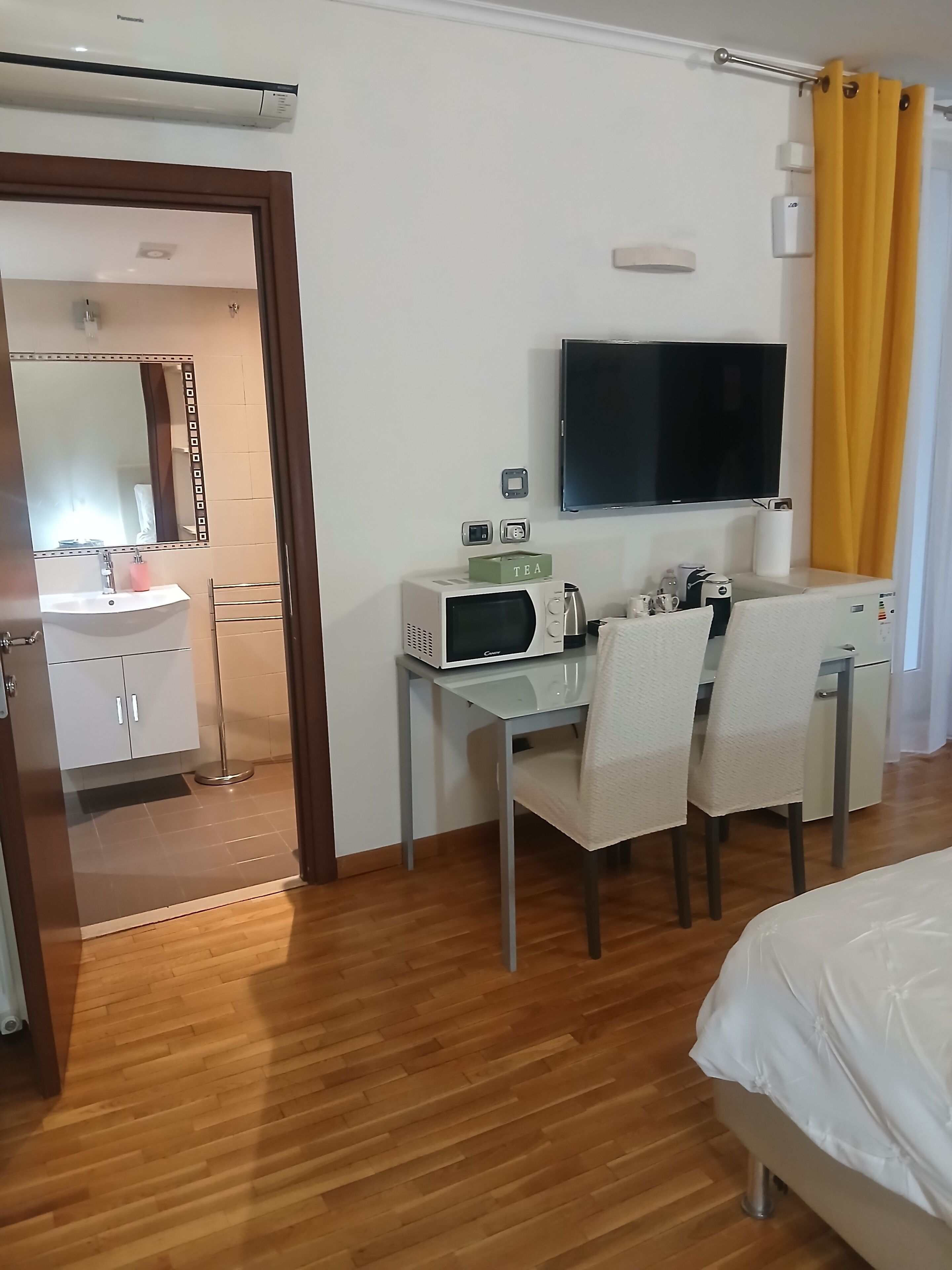 A hotel room with a bed, a desk with chairs, a microwave, a television, and a bathroom visible through an open door.