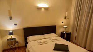 Un letto rifatto con lenzuola bianche e un asciugamano piegato, una lampada da comodino, un piccolo tavolino con lampada e un punto luce a parete.