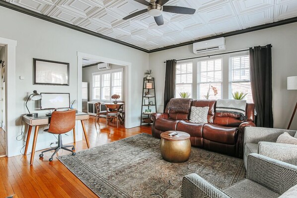A spacious living room with a leather sofa, a coffee table, and a desk with a chair. There is a ceiling fan, a bookshelf, and a window with curtains.