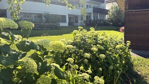Ein modernes Gebäude mit flachem Dach und großen Fenstern, umgeben von einer gepflegten Rasenfläche und blühenden Pflanzen.
