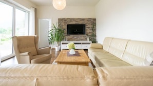 Ein Wohnzimmer mit einem beigefarbenen Sofa, einem Holztisch, einem beigefarbenen Sessel und einem an einer Steinwand montierten Fernseher.