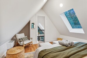 A modern, well-lit attic bedroom with a sloped ceiling, a large skylight, a bed with a green blanket, a wicker chair, and a view of the outdoors. - Rocky Falls | Hocking Hills, Modern Cabin, Hot Tub (Rockbridge)