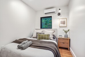A bedroom with a bed, a nightstand, a window with a forest view, an air conditioner, and a framed picture on the wall.