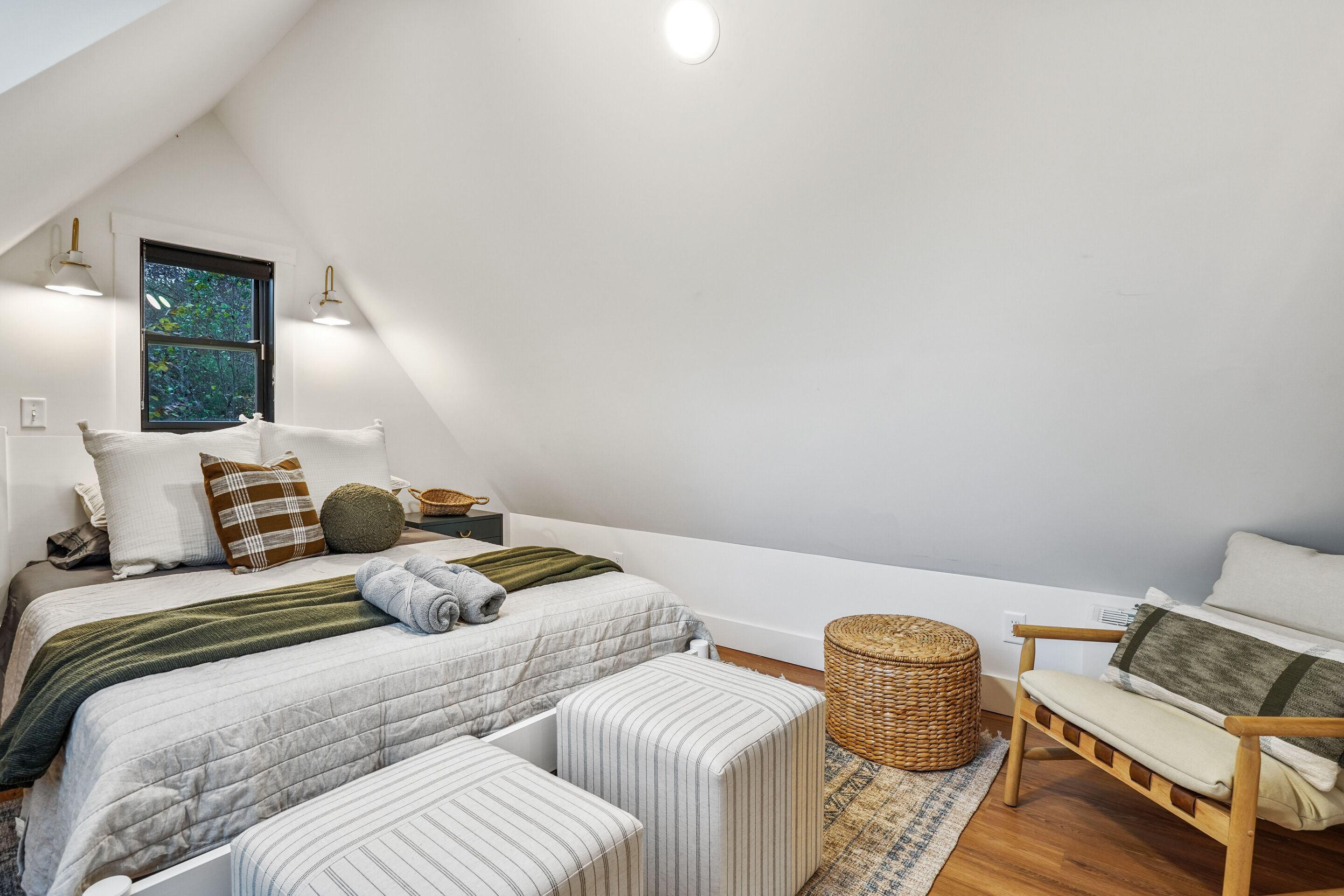 Un dormitorio moderno y bien iluminado, con techo inclinado, un ventanal grande, una cama con varias almohadas, una mesita de noche, una silla y una cesta tejida.