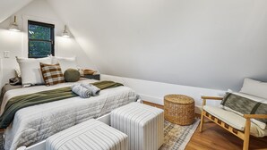 Ein modernes, helles Schlafzimmer mit schrägem Dach, einem großen Fenster, einem Bett mit vielen Kissen, einem Nachttisch, einem Sessel und einem gewebten Korb.